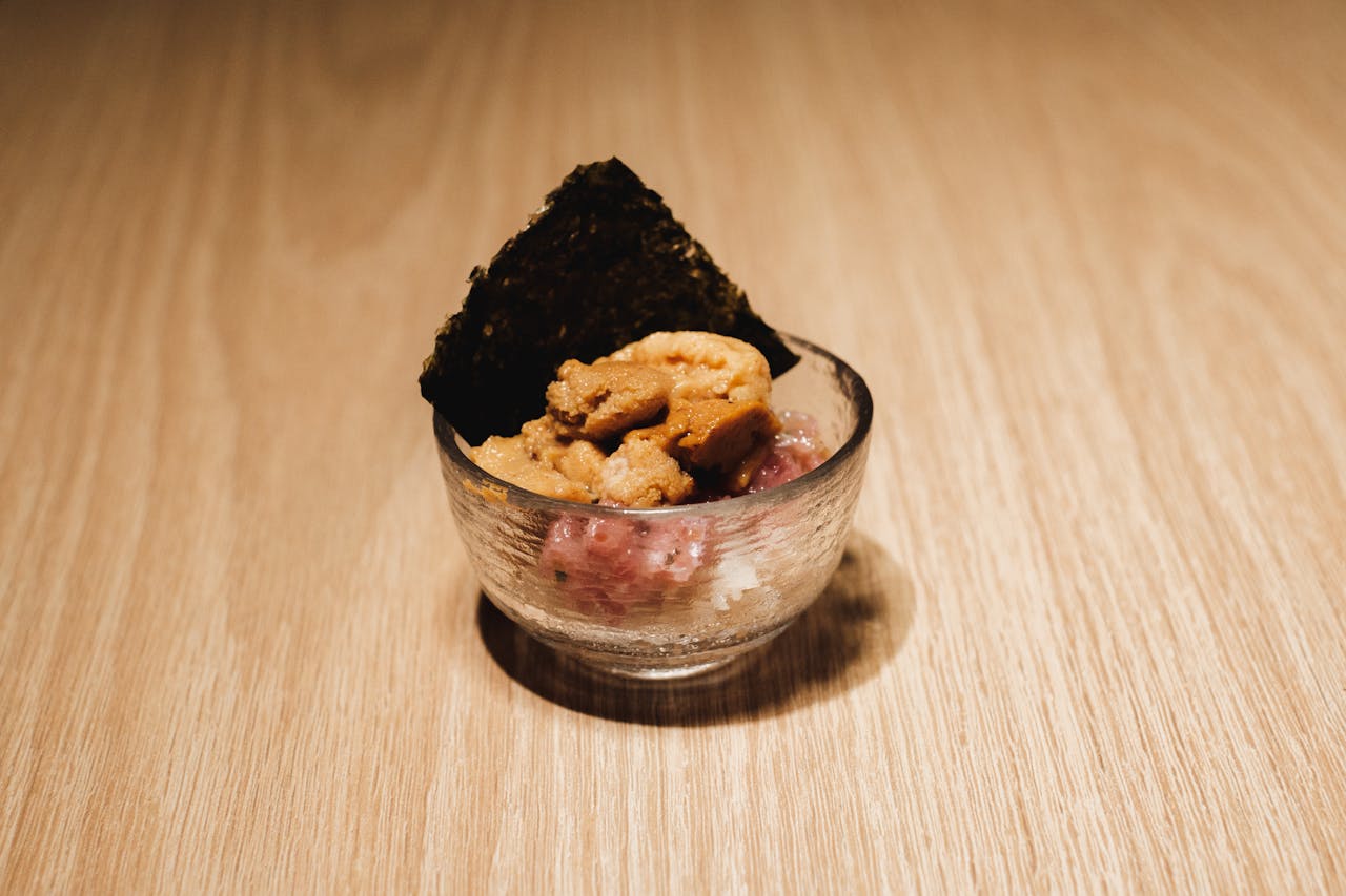 Delicious uni and tuna tartare served with nori in a clear glass bowl on a wooden surface.