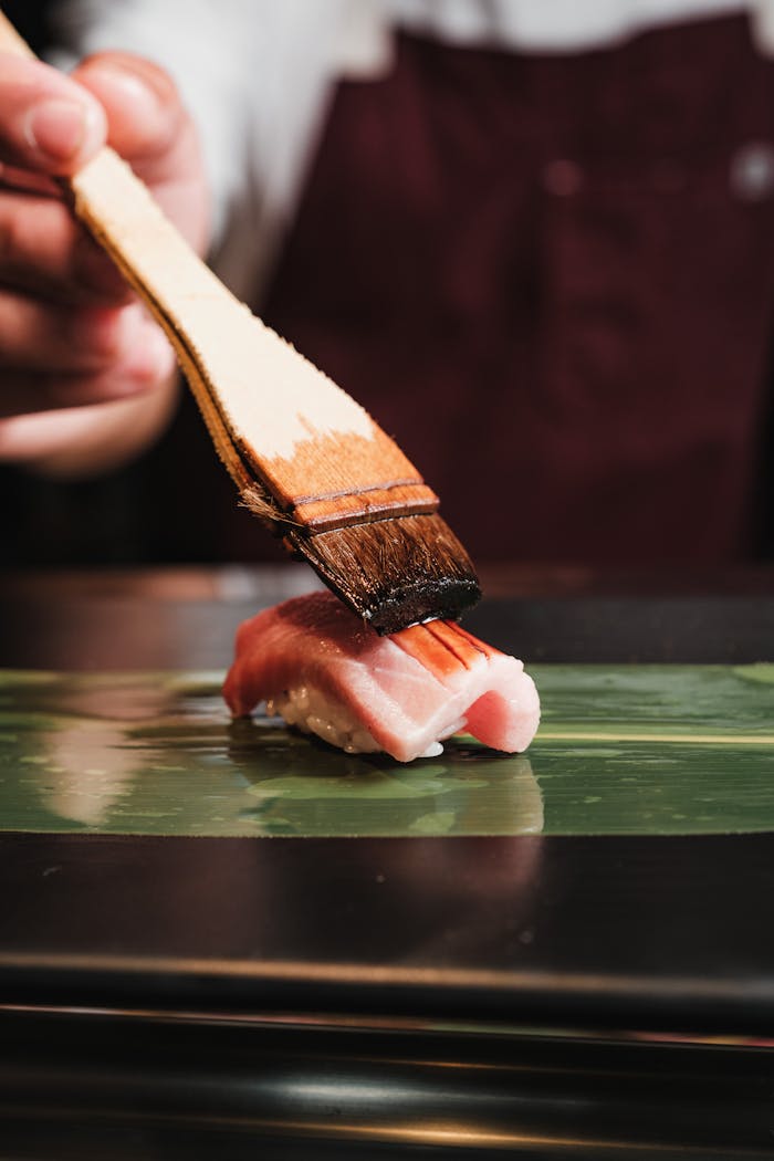 Close-up of a chef using a brush to glaze a piece of sushi nigiri on a leaf, showcasing Japanese culinary art.